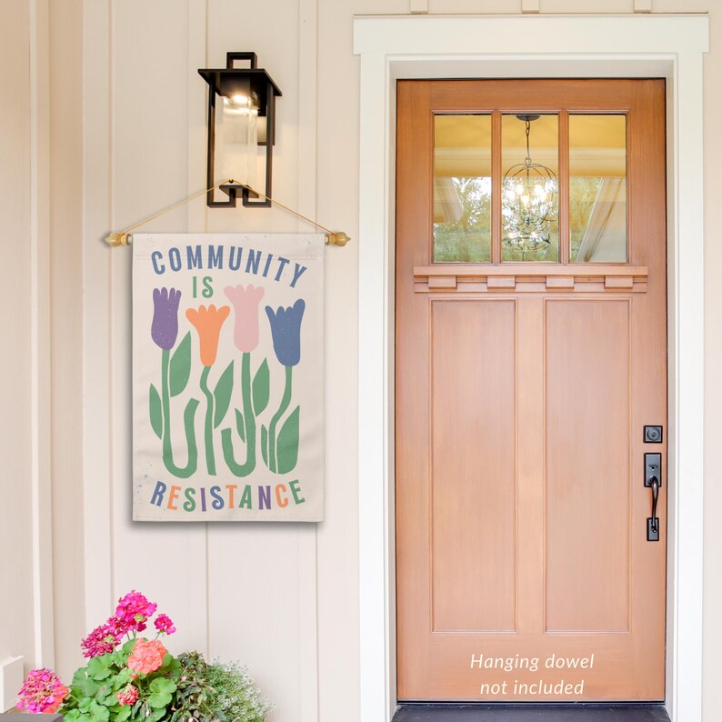 May include: A beige canvas flag with the text "Community is Resistance" in black lettering. The flag features a stylised design of four colourful tulips with green stems and leaves.