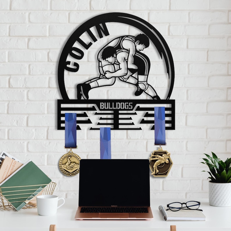 Op de afbeelding: Zwarte metalen worstelmedaillehouder met de naam "COLIN" en het woord "BULLDOGS". De houder toont twee worstelaars en heeft sleuven voor medailles met blauwe linten. Twee gouden medailles hangen aan de houder.