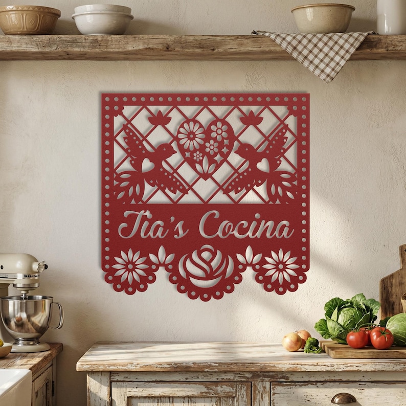 Op de afbeelding: Rode metalen wanddecoratie met een decoratief ontwerp. Het ontwerp omvat twee vogels, een hart met bloemendetails en de tekst "Tia's Cocina". De onderste rand heeft een geschulpte bloemenpatroon.