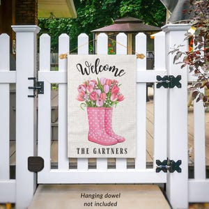 May include: A welcome garden flag with a pink and white floral design in pink rain boots. The text "Welcome" and "The Gartners" is printed on the flag.