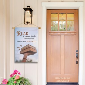 Może przedstawiać: Dekoracyjna flaga zewnętrzna z niebieskim tłem i brązowym nadrukiem tekstu "READ Banned Books Free Societies READ FREELY". Flaga przedstawia rękę trzymającą w powietrzu stos książek.