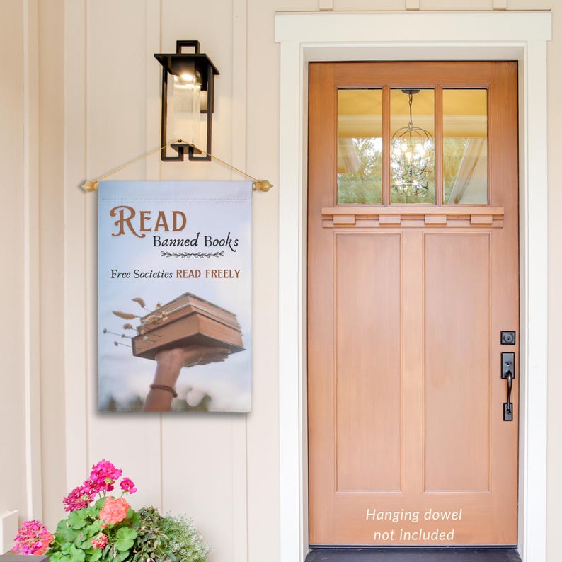 Może przedstawiać: Dekoracyjna flaga zewnętrzna z niebieskim tłem i brązowym nadrukiem tekstu "READ Banned Books Free Societies READ FREELY". Flaga przedstawia rękę trzymającą w powietrzu stos książek.