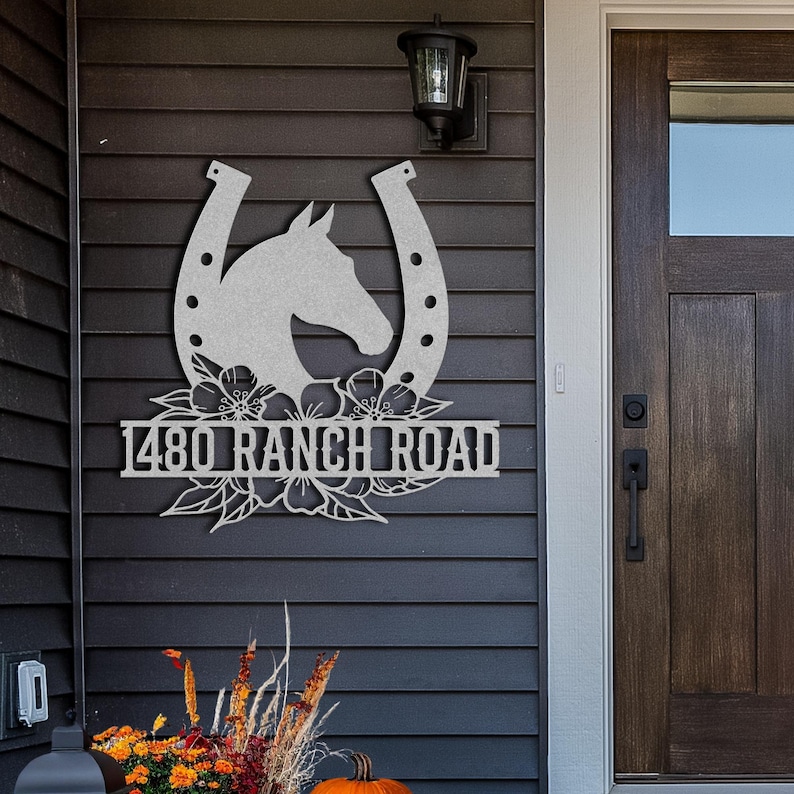 May include: A silver metal sign featuring a horse head within a horseshoe design, with floral accents and the text "1480 RANCH ROAD". The sign is mounted on a dark gray exterior wall near a wooden door.
