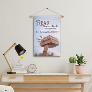 Może przedstawiać: Biały baner z pł&oacute;tna z tekstem "READ Banned Books" i "Free Societies READ FREELY". Ręka trzyma stos książek w g&oacute;rze na tle niebieskiego nieba.