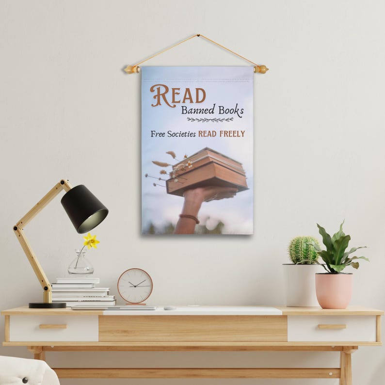 Może przedstawiać: Biały baner z pł&oacute;tna z tekstem "READ Banned Books" i "Free Societies READ FREELY". Ręka trzyma stos książek w g&oacute;rze na tle niebieskiego nieba.