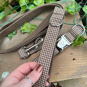 May include: A brown and white gingham dog collar and leash set. The collar has a silver buckle and a silver D-ring. The leash has a silver clip.