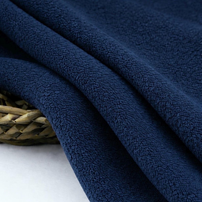 May include: Close-up of a navy blue fleece fabric. The fabric is soft and textured, with a slightly uneven surface. The folds and shadows create depth and dimension, highlighting the plush texture of the material. The fabric is likely used for clothing or blankets.