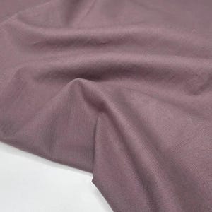 Puede incluir: Primer plano de una tela malva con textura y pliegues visibles. La tela tiene un aspecto suave y mate y un brillo sutil. El color es un rosa apagado y polvoriento, con un ligero matiz p&uacute;rpura.