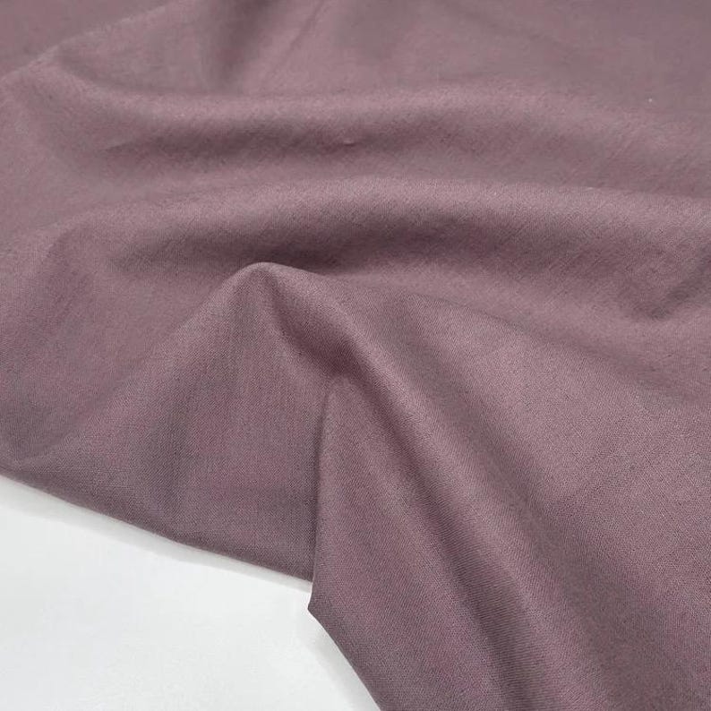 Puede incluir: Primer plano de una tela malva con textura y pliegues visibles. La tela tiene un aspecto suave y mate y un brillo sutil. El color es un rosa apagado y polvoriento, con un ligero matiz p&uacute;rpura.