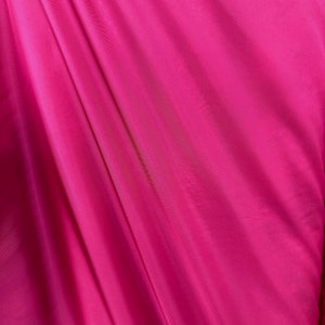 Puede incluir: Primer plano de una tela lisa de color rosa con un ligero brillo. La tela est&aacute; drapeada y doblada, creando l&iacute;neas suaves y fluidas.