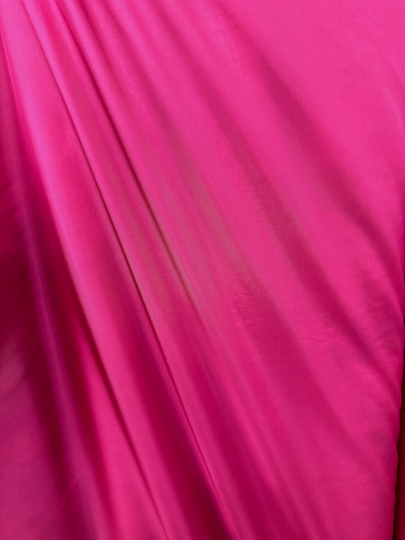 Puede incluir: Primer plano de una tela lisa de color rosa con un ligero brillo. La tela est&aacute; drapeada y doblada, creando l&iacute;neas suaves y fluidas.