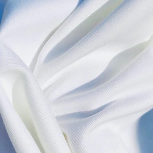 May include: Close-up of white fabric with visible texture and folds. The fabric is brightly lit, creating shadows that emphasize the texture and flow of the material. The overall impression is of a soft, flowing textile.