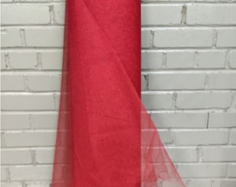 May include: A roll of red glitter tulle fabric. The fabric is folded and standing upright on a table. The fabric is in front of a white brick wall.