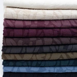 May include: A stack of folded fabric pieces in various colours including white, cream, plum, dark purple, brown, charcoal, olive, teal, light blue, and black. Each piece of fabric features a consistent, repeating tonal leaf pattern.