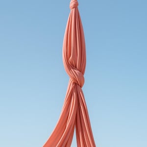 May include: A length of coral-coloured aerial silk is knotted in the middle, with the ends cascading downwards. The fabric is set against a clear, light blue sky. The image highlights the texture and flow of the fabric.
