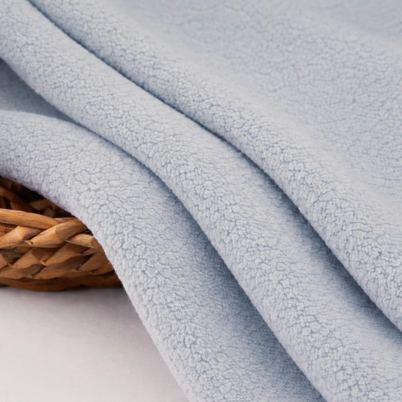 May include: Close-up of a light blue fleece fabric with a soft, textured surface. The fabric is folded in gentle waves, revealing its plush texture. The background includes a woven basket.