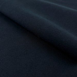 May include: Close-up of a dark navy blue fabric with a subtle texture. The fabric is folded, creating shadows and highlighting the material's weave. The overall appearance suggests a soft, possibly knitted, textile.