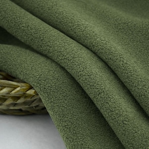 May include: Close-up of olive green fleece fabric with a soft, textured surface. The fabric is draped in gentle folds, creating shadows and highlighting the plush texture. The material appears thick and warm, suitable for various crafting projects.