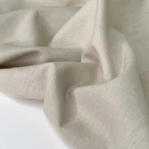 Puede incluir: Primer plano de una tela de lino beige con un aspecto suave y texturizado. La tela est&aacute; drapeada, mostrando pliegues y sombras. El color neutro y la textura natural sugieren un enfoque en textiles y decoraci&oacute;n del hogar.
