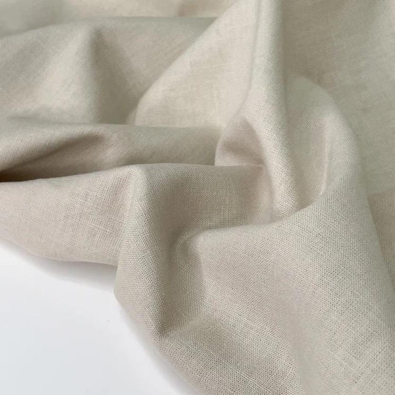 Puede incluir: Primer plano de una tela de lino beige con un aspecto suave y texturizado. La tela est&aacute; drapeada, mostrando pliegues y sombras. El color neutro y la textura natural sugieren un enfoque en textiles y decoraci&oacute;n del hogar.