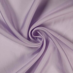 May include: A close-up shot of a lavender-colored fabric with a silky sheen. The fabric is draped and swirled, creating soft folds and shadows. The material appears smooth and luxurious, with a subtle sheen that catches the light.