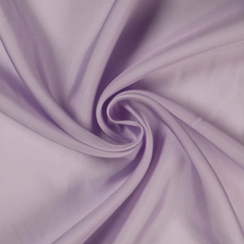 May include: A close-up shot of a lavender-colored fabric with a silky sheen. The fabric is draped and swirled, creating soft folds and shadows. The material appears smooth and luxurious, with a subtle sheen that catches the light.