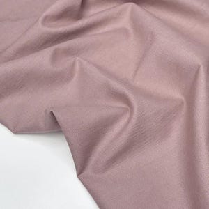 Puede incluir: Primer plano de una tela rosa empolvado con textura visible y pliegues. La tela es de color liso y parece ser un material de peso medio, adecuado para proyectos de ropa o decoraci&oacute;n del hogar. La iluminaci&oacute;n resalta las sutiles variaciones en la superficie de la tela.