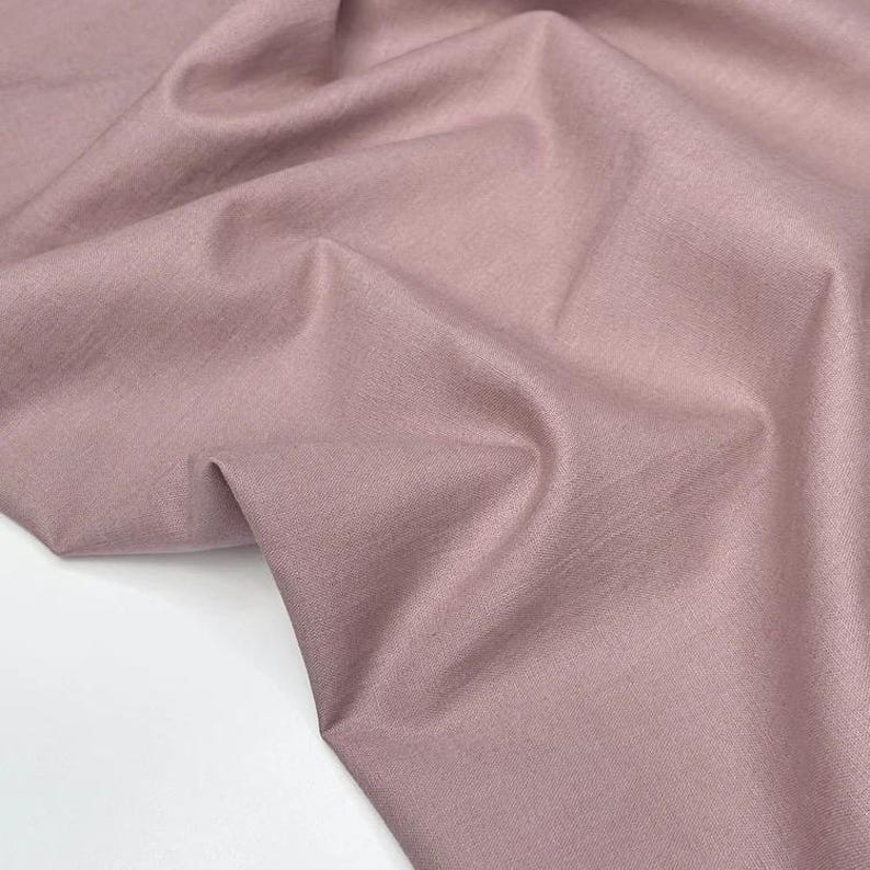 Puede incluir: Primer plano de una tela rosa empolvado con textura visible y pliegues. La tela es de color liso y parece ser un material de peso medio, adecuado para proyectos de ropa o decoraci&oacute;n del hogar. La iluminaci&oacute;n resalta las sutiles variaciones en la superficie de la tela.