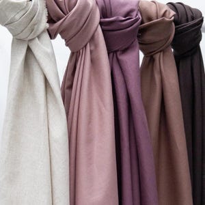 Puede incluir: Colecci&oacute;n de cinco bufandas en varios colores: beige, malva, morado, marr&oacute;n y marr&oacute;n oscuro. Las bufandas est&aacute;n colgadas de una barra, mostrando su textura suave y variaciones de color. La tela parece ligera y adecuada para varias estaciones.