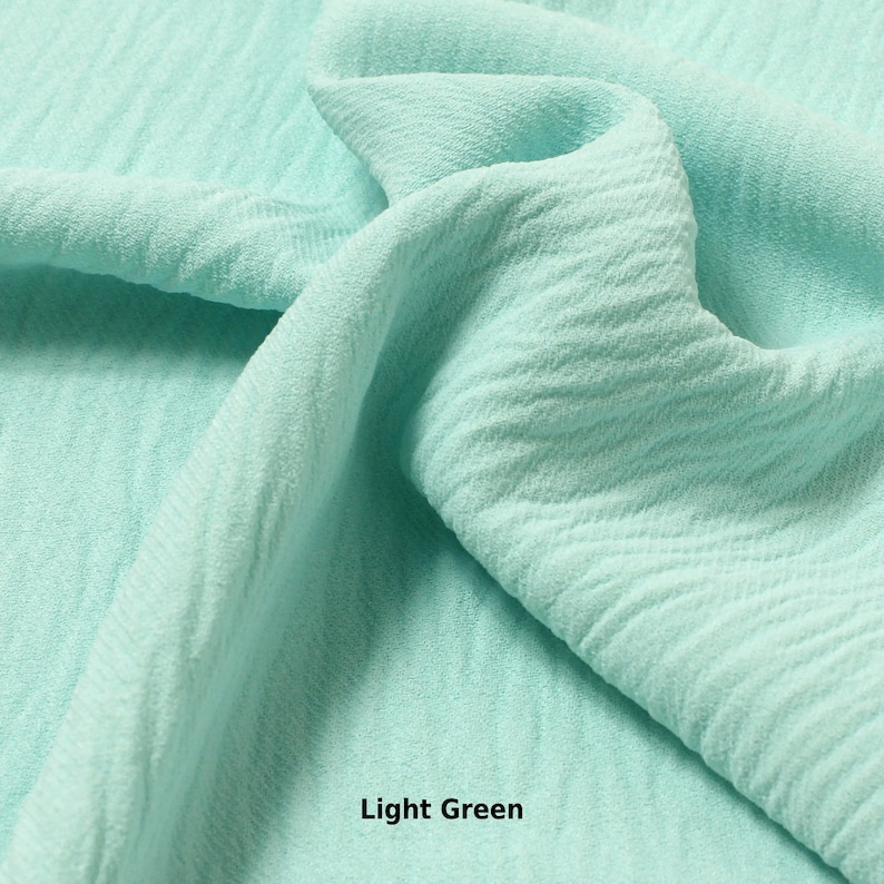 May include: A close-up of a light green fabric with a textured surface. The fabric is folded and draped, showing its soft and flowing texture.