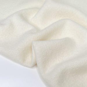 Puede incluir: Primer plano de una tela suave de color blanco roto con una superficie ligeramente texturizada. La tela está drapeada y doblada, creando sombras y profundidad. El material parece cálido y acogedor, posiblemente lana o forro polar.
