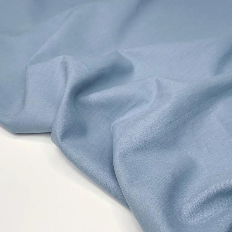 Puede incluir: Primer plano de una tela azul claro con pliegues suaves y un ligero brillo. La tela parece ligera, posiblemente lino o similar, y se drapea de forma natural. El color es un azul pastel suave.