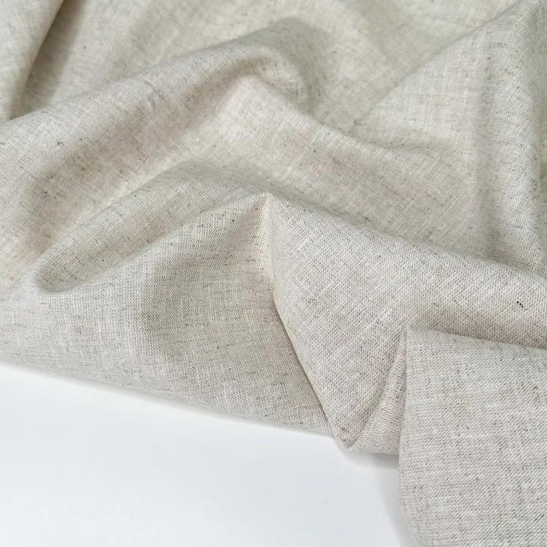 Puede incluir: Primer plano de una tela de lino beige claro doblada. La tela tiene un aspecto texturizado con sutiles variaciones de color, creando un aspecto natural y org&aacute;nico. Los pliegues y las sombras a&ntilde;aden profundidad y dimensi&oacute;n al textil.