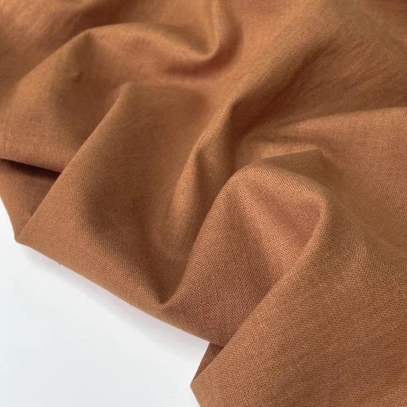Puede incluir: Primer plano de un trozo de tela marr&oacute;n doblada. La tela tiene un aspecto texturizado con pliegues y arrugas visibles. El color es un tono c&aacute;lido y terroso, con luces y sombras que a&ntilde;aden profundidad al material.