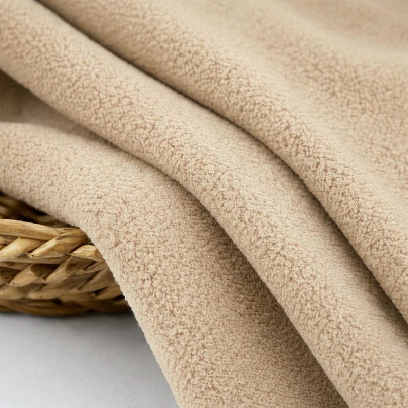 May include: Close-up of a tan fleece fabric with a soft, textured surface. The fabric is draped in gentle folds, revealing its plush texture. The neutral color and cozy appearance suggest warmth and comfort, suitable for various sewing projects.