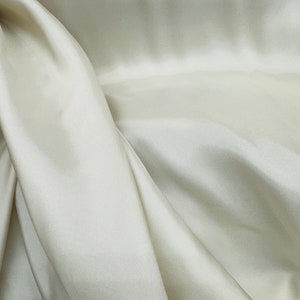 Puede incluir: Primer plano de una tela lisa de color blanco roto con pliegues y arrugas suaves. La tela tiene un brillo sutil y una sensaci&oacute;n de lujo.