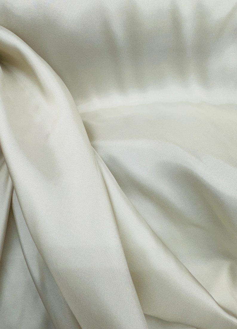 Puede incluir: Primer plano de una tela lisa de color blanco roto con pliegues y arrugas suaves. La tela tiene un brillo sutil y una sensaci&oacute;n de lujo.
