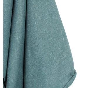 May include: Close-up of teal fabric, showing a textured surface with lighter flecks. The edge is folded over, forming a rounded hem. The fabric appears soft and flowing. The colour is a muted teal, suggesting a garment or textile.
