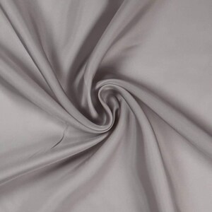 May include: A close-up shot of a swirled, light gray fabric. The fabric has a smooth, silky texture with soft folds and subtle highlights. The material appears to be a solid color, with no visible patterns or designs.