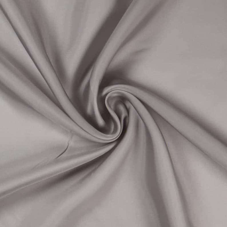 May include: A close-up shot of a swirled, light gray fabric. The fabric has a smooth, silky texture with soft folds and subtle highlights. The material appears to be a solid color, with no visible patterns or designs.