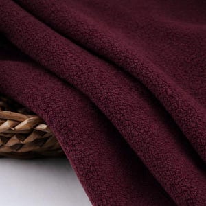 May include: Close-up of a burgundy fleece fabric with a soft, textured surface. The fabric is draped in folds, revealing its plush texture and rich color. A woven basket is partially visible in the lower left corner.