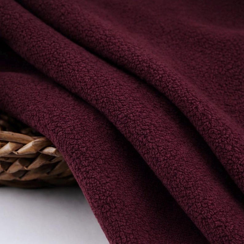 May include: Close-up of a burgundy fleece fabric with a soft, textured surface. The fabric is draped in folds, revealing its plush texture and rich color. A woven basket is partially visible in the lower left corner.