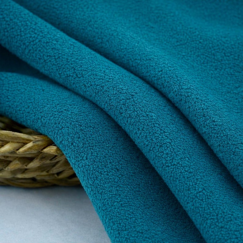 May include: Close-up of a teal fleece fabric with a soft, textured surface. The fabric is draped in gentle folds, revealing its plush texture. A woven basket is partially visible in the lower left corner, adding a touch of natural contrast.