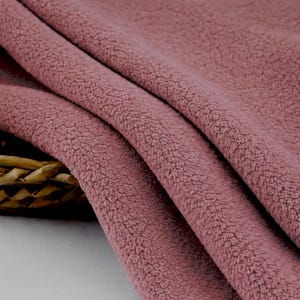 May include: Close-up of a folded piece of dusty rose-colored fabric. The material has a soft, textured appearance, with a dense, looped pile. The fabric is draped, showing the folds and depth of the material. The texture suggests a cozy, warm textile.