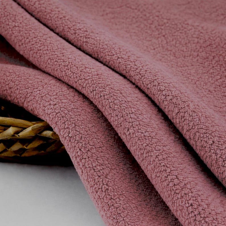 May include: Close-up of a folded piece of dusty rose-colored fabric. The material has a soft, textured appearance, with a dense, looped pile. The fabric is draped, showing the folds and depth of the material. The texture suggests a cozy, warm textile.