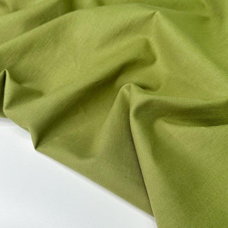 Puede incluir: Primer plano de una tela verde oliva doblada. La tela tiene un aspecto ligeramente texturizado y est&aacute; drapeada en pliegues suaves. El color es un verde apagado y terroso, con sutiles variaciones de tono debido a los pliegues y la iluminaci&oacute;n. La tela parece ser de peso medio.