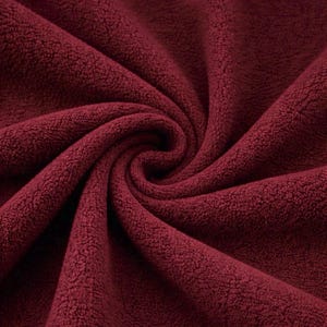 May include: Close-up of a deep burgundy fleece fabric, tightly swirled to reveal its texture. The fabric has a soft, plush appearance, with a dense, fuzzy surface. The color is a rich, dark red, with subtle variations in tone.