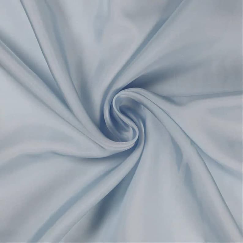 May include: A close-up shot of a light blue fabric with a silky texture. The fabric is swirled, creating soft folds and shadows. The color is a pale, muted blue, and the fabric appears to be lightweight and flowing.