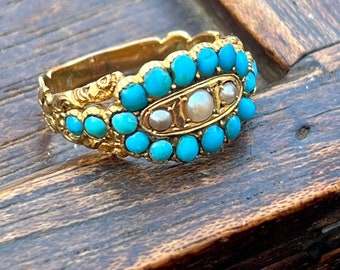 May include: A gold ring featuring a band of small, round, turquoise stones. The ring has a central oval design with three pearls. The ring is set on a wooden surface.