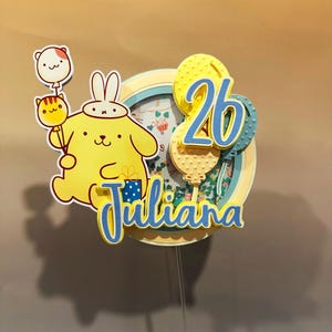 Puede incluir: Adorno para tarta de cumpleaños amarillo y azul con un personaje de dibujos animados sosteniendo globos. El adorno incluye el número "26" y el nombre "Juliana" en escritura azul. El diseño está superpuesto con un fondo floral.